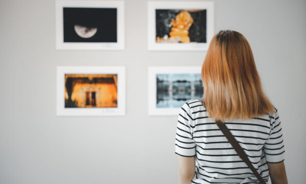 Asian young woman standing she looking art gallery in front of colorful framed paintings pictures on white wall, female watch at photo frame to leaning against at exhibit museum, Back view