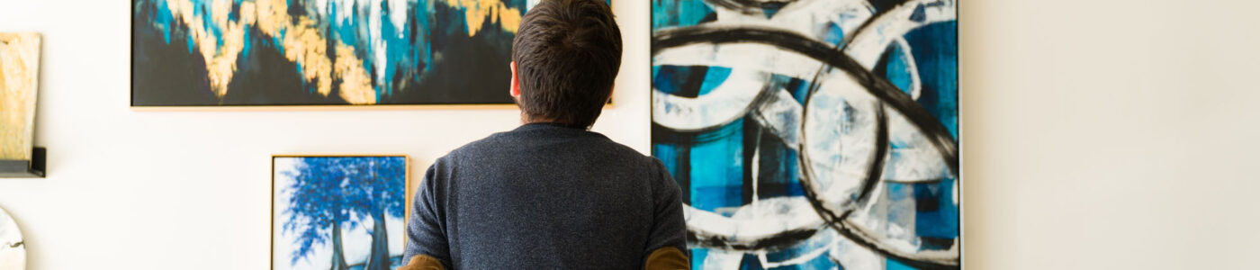 Hispanic male visitor looking reflective while sitting on a bench and admiring the various paintings on the wall of an art gallery